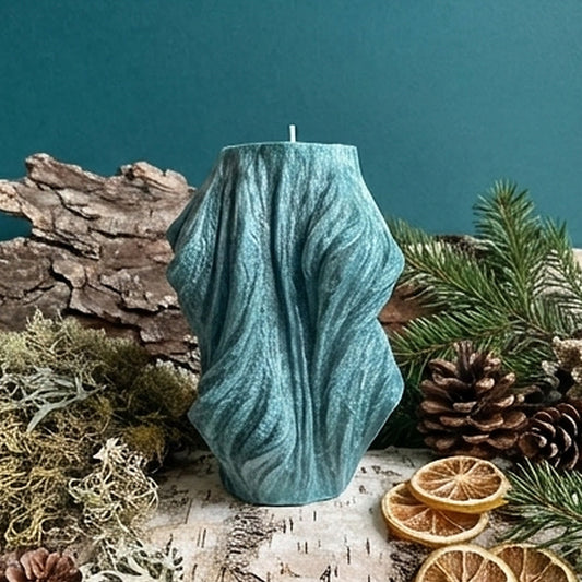 Teal textured candle on a wooden log with pine cones, dried oranges, and greenery against a teal background
