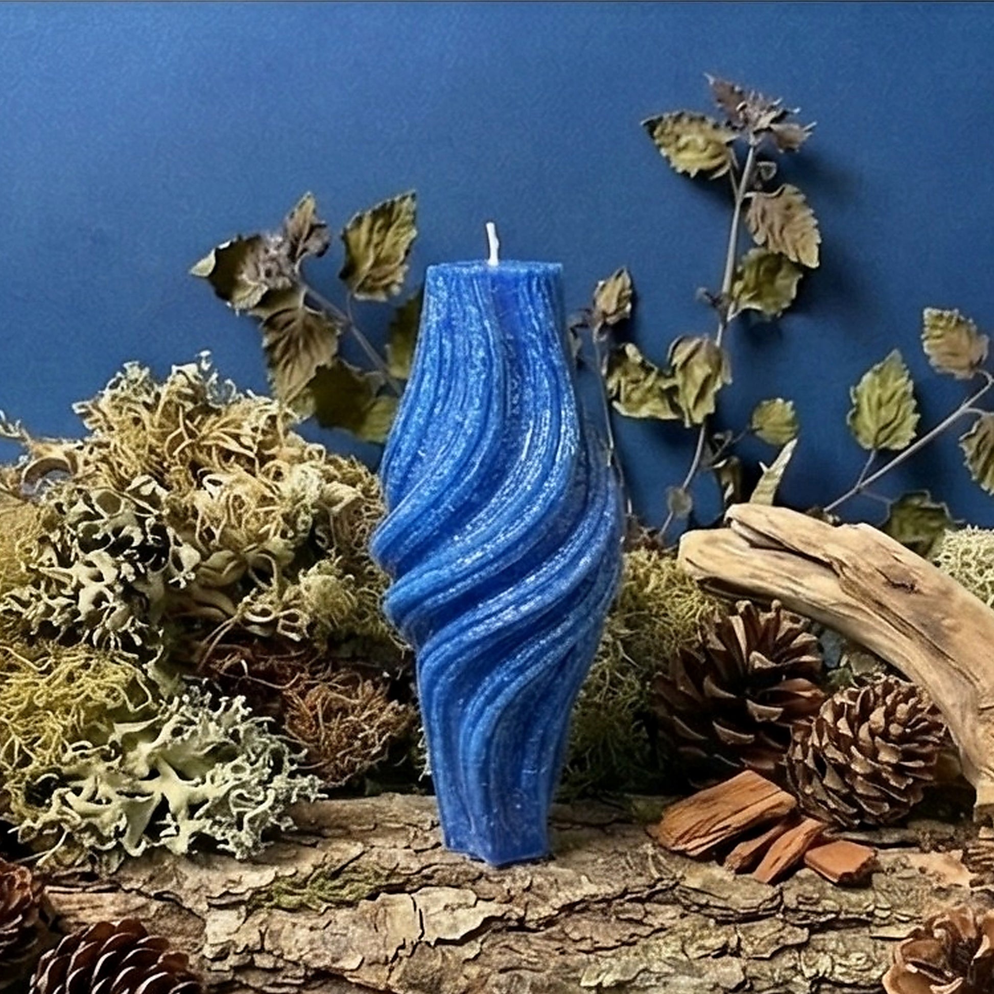 Blue candle with wavy texture on a natural log surrounded by pine cones, wood chips, and foliage against a blue background.