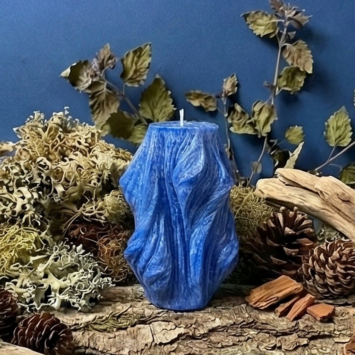 Blue candle on a natural setting with wood, moss, and pinecones against a blue background