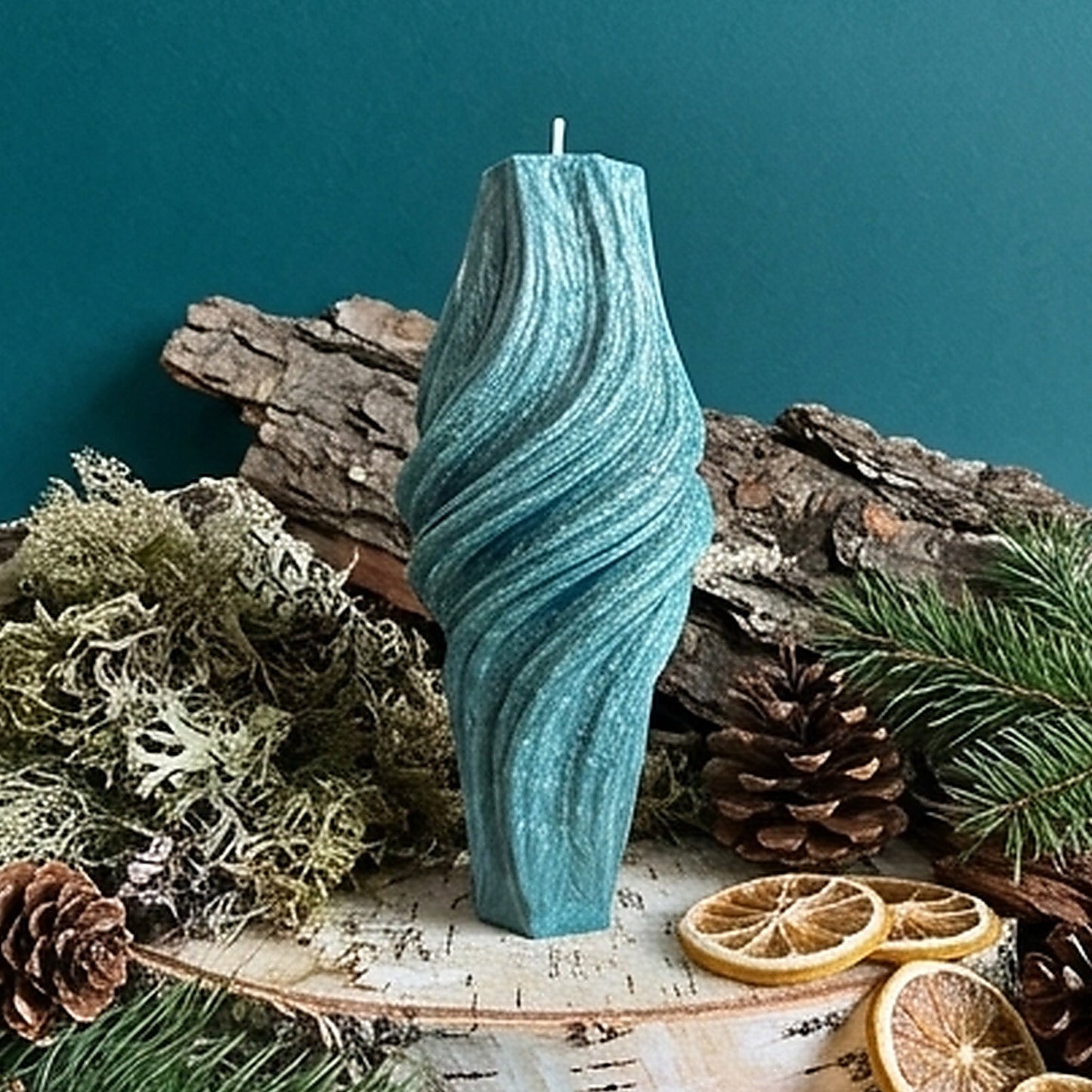 Teal textured candle on a rustic wooden surface with pine cones and dried oranges against a teal background